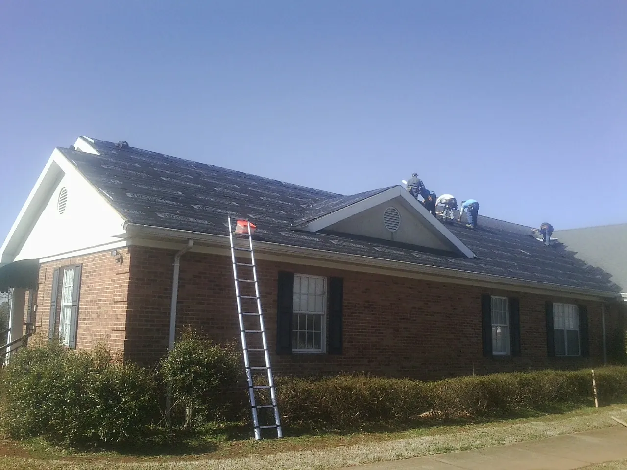 Crew tearing shingles off a brick ranch roof with a ladder set — Emergency Roof Repair work by our crew in East China